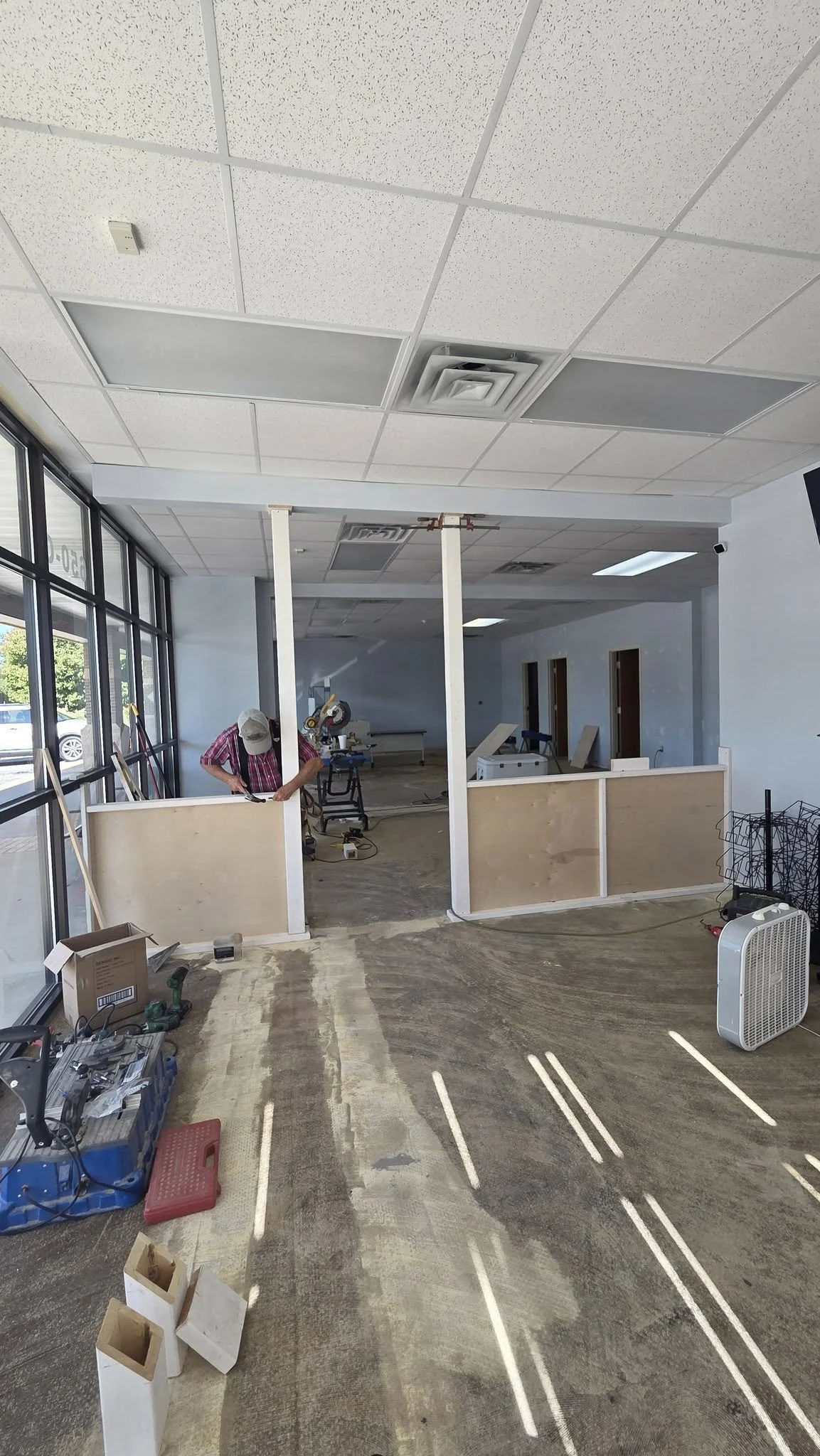 Interior of a building under renovation, with drywall being installed, construction tools on the floor, and a person working.