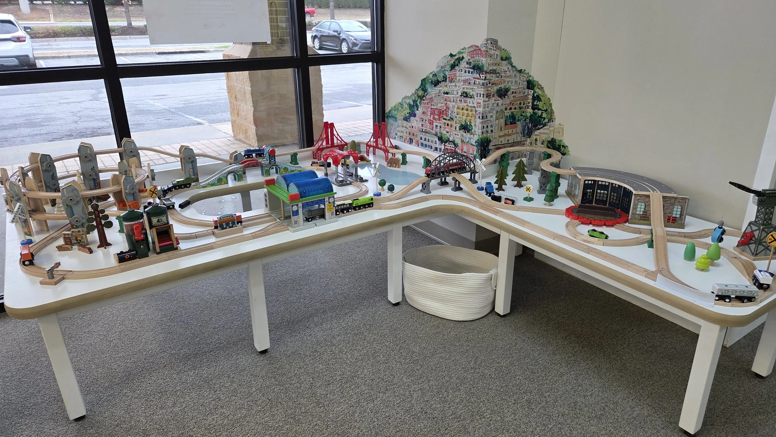 A large wooden model train set on a white table next to a window, featuring tracks, bridges, buildings, trees, and miniature trains, with a painted hillside mural on the wall in the background.