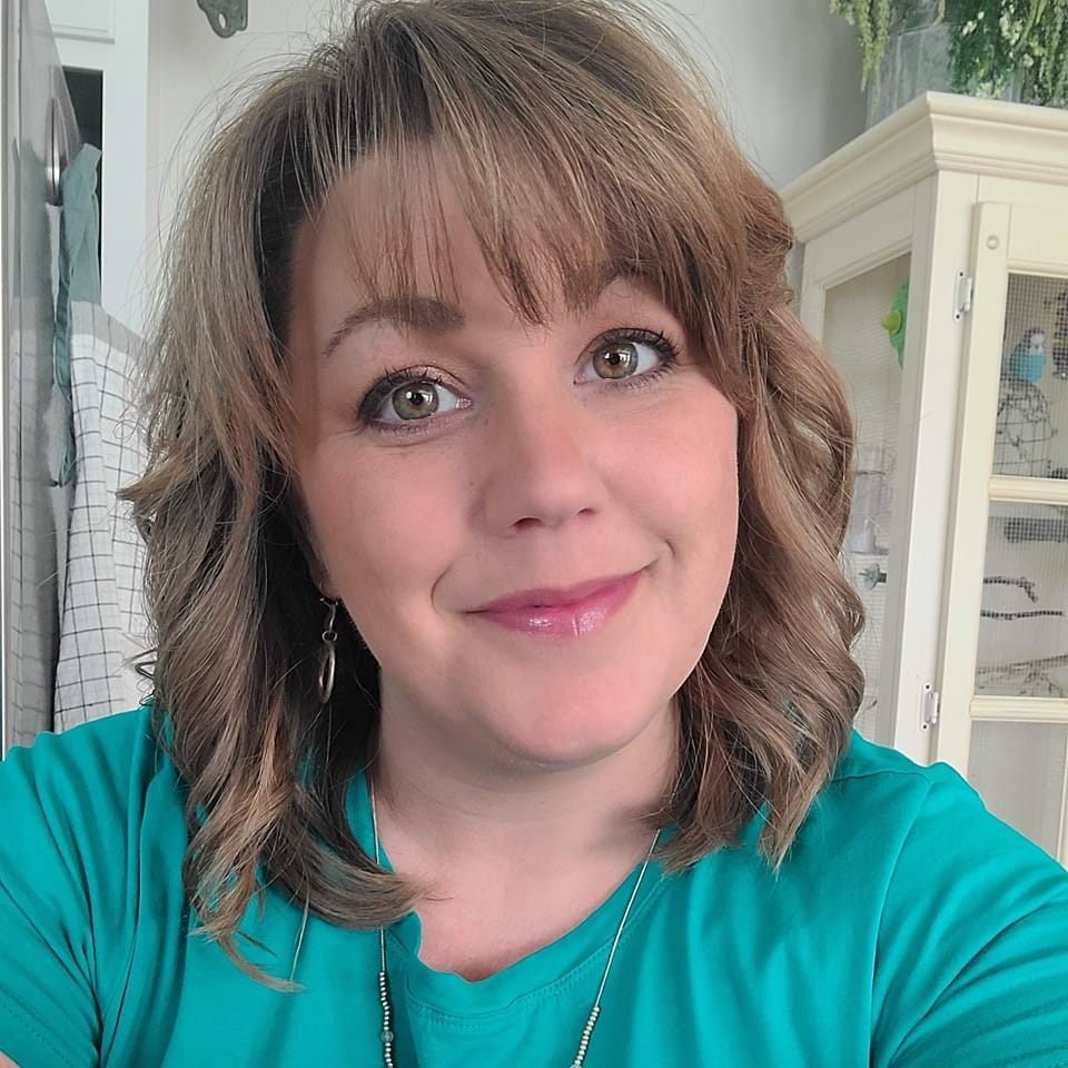 A woman with shoulder-length wavy brown hair and hazel eyes, smiling at the camera, wearing a teal shirt, with a white cabinet and some greenery in the background.