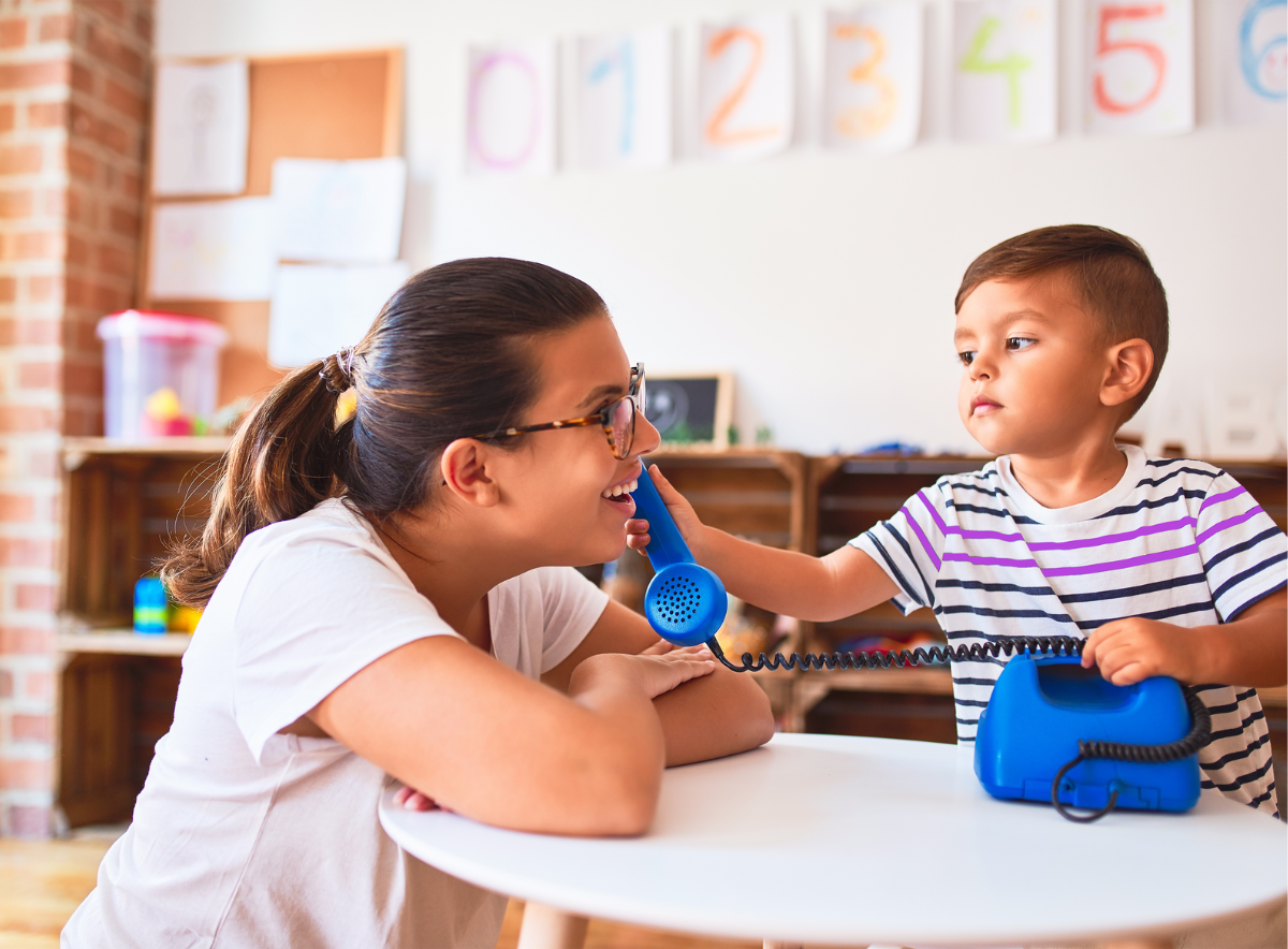A young boy offers a toy phone to a woman, pretending to talk on it, in a classroom setting.