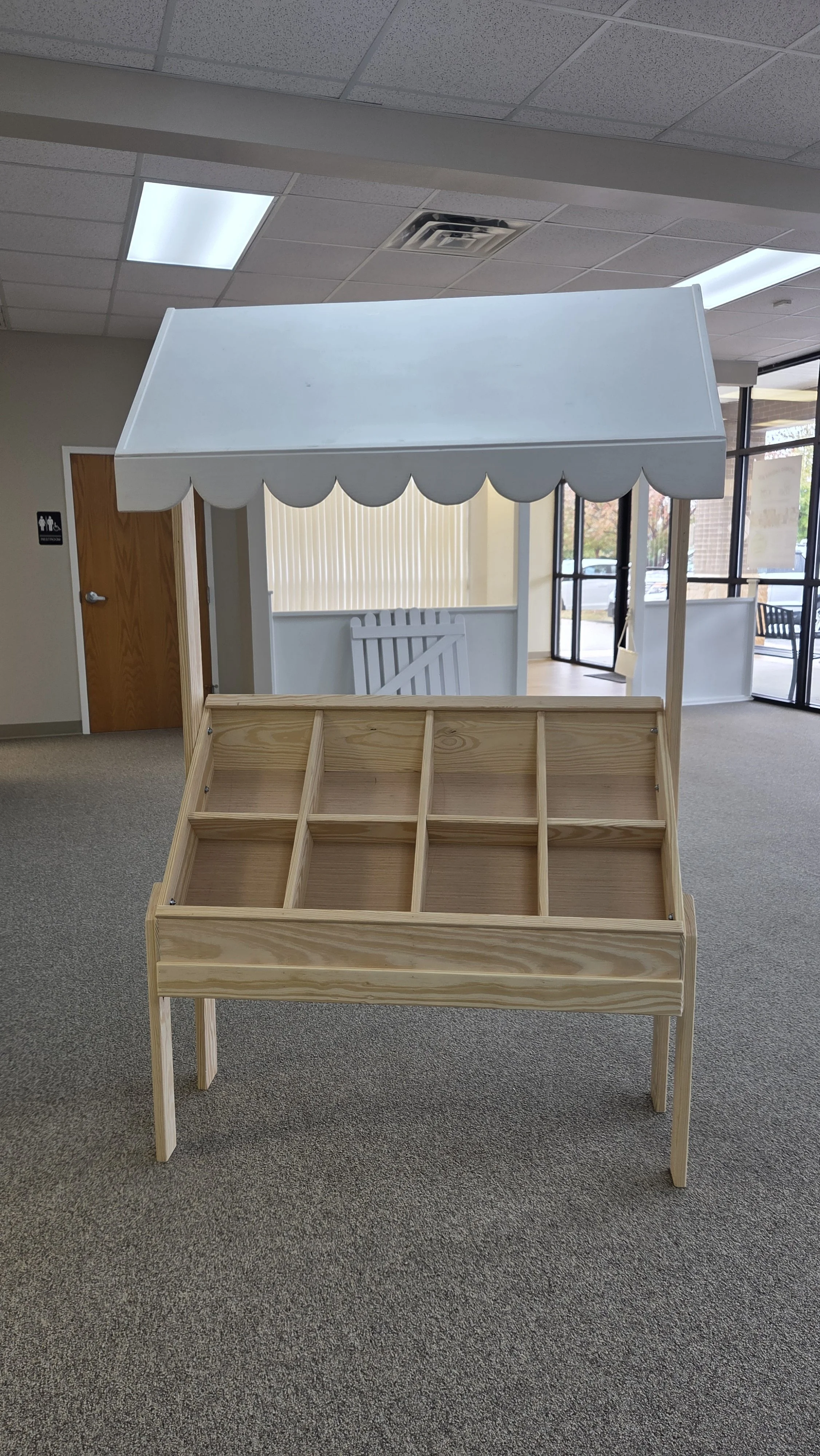 A small wooden display stand with multiple compartments and a white canopy, resembling a market or ice cream stand, situated indoors.