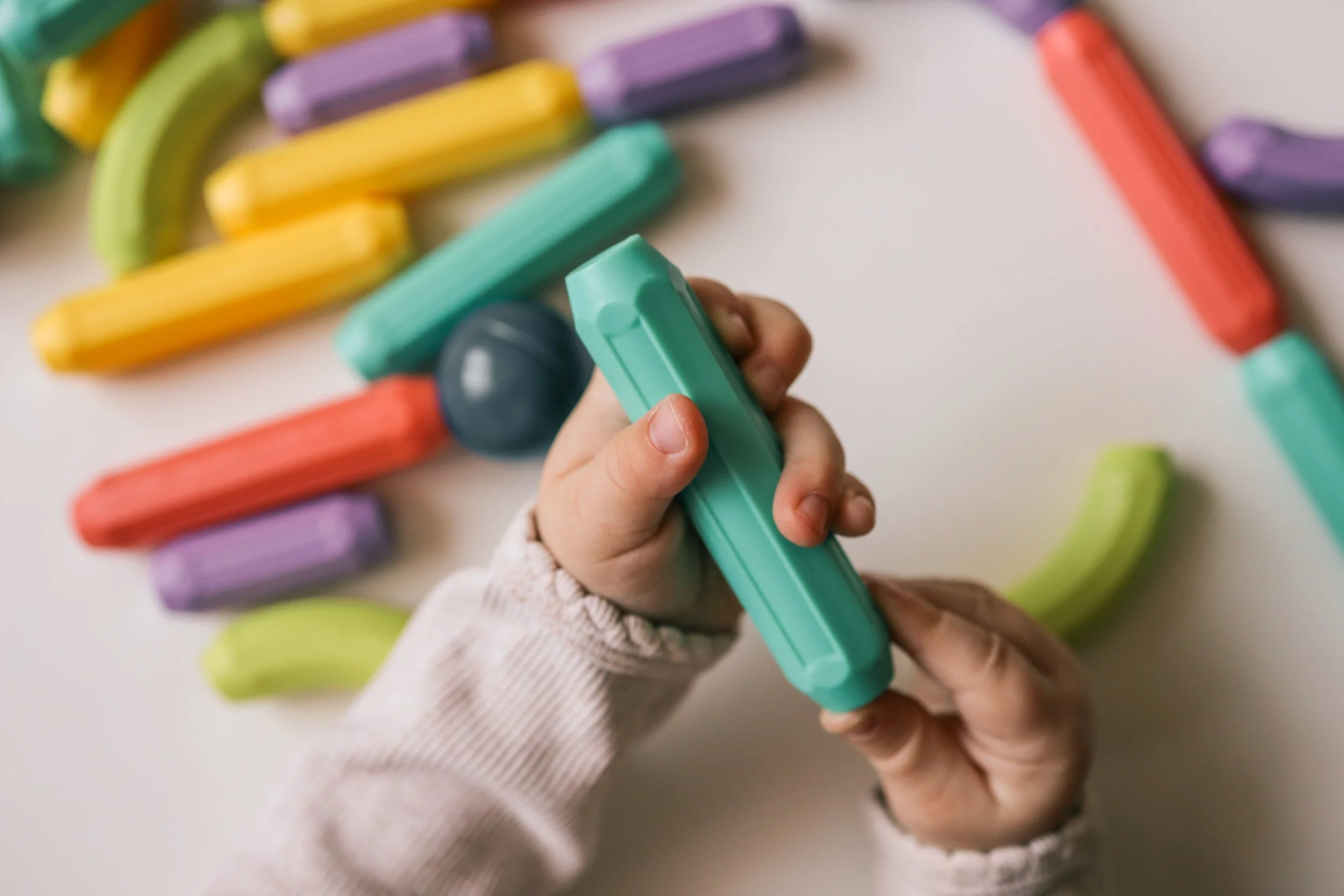 Child's hands holding a piece of colorful chalk, with a rainbow made of chalk pieces on a white surface.