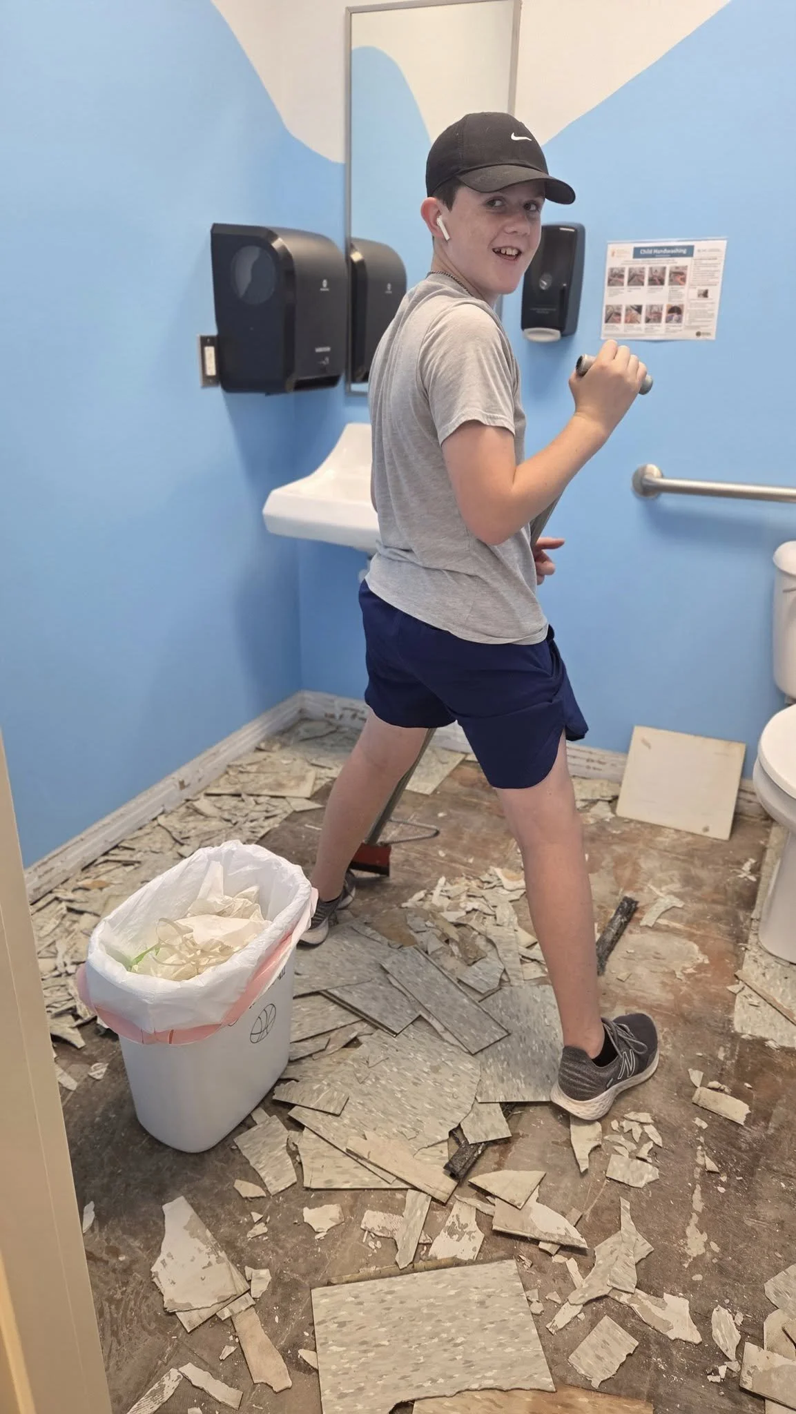 A young boy in a bathroom with light blue walls, black Nike cap, gray t-shirt, navy shorts, and gray sneakers, standing on a floor covered with broken tiles, holding a tool, with a trash can nearby, and bathroom fixtures including a sink and toilet v