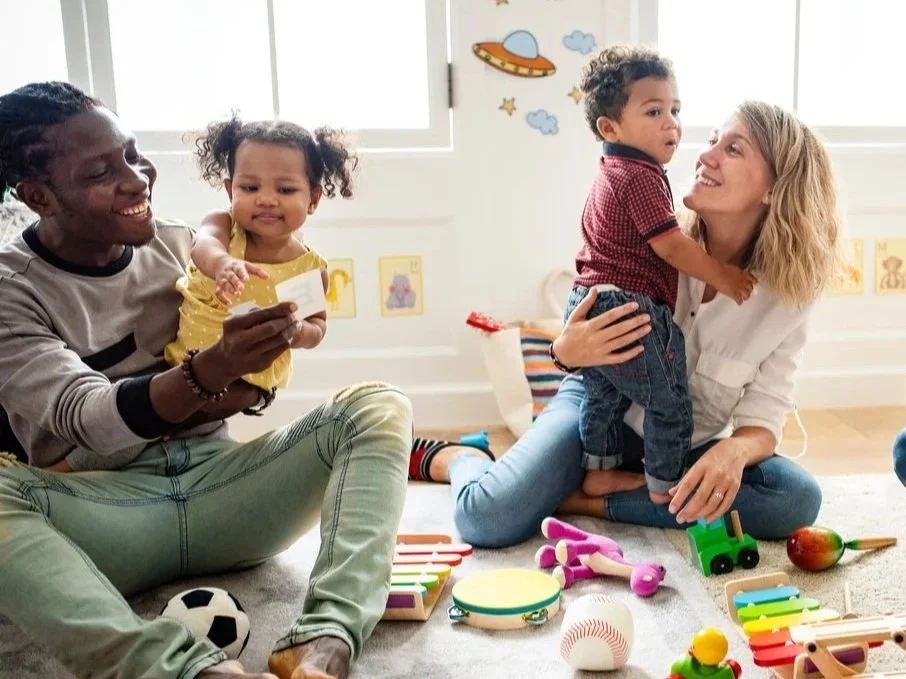 Two adults, a man and a woman, with two young children, playing with toys in a bright, cheerful room with space-themed wall decorations.