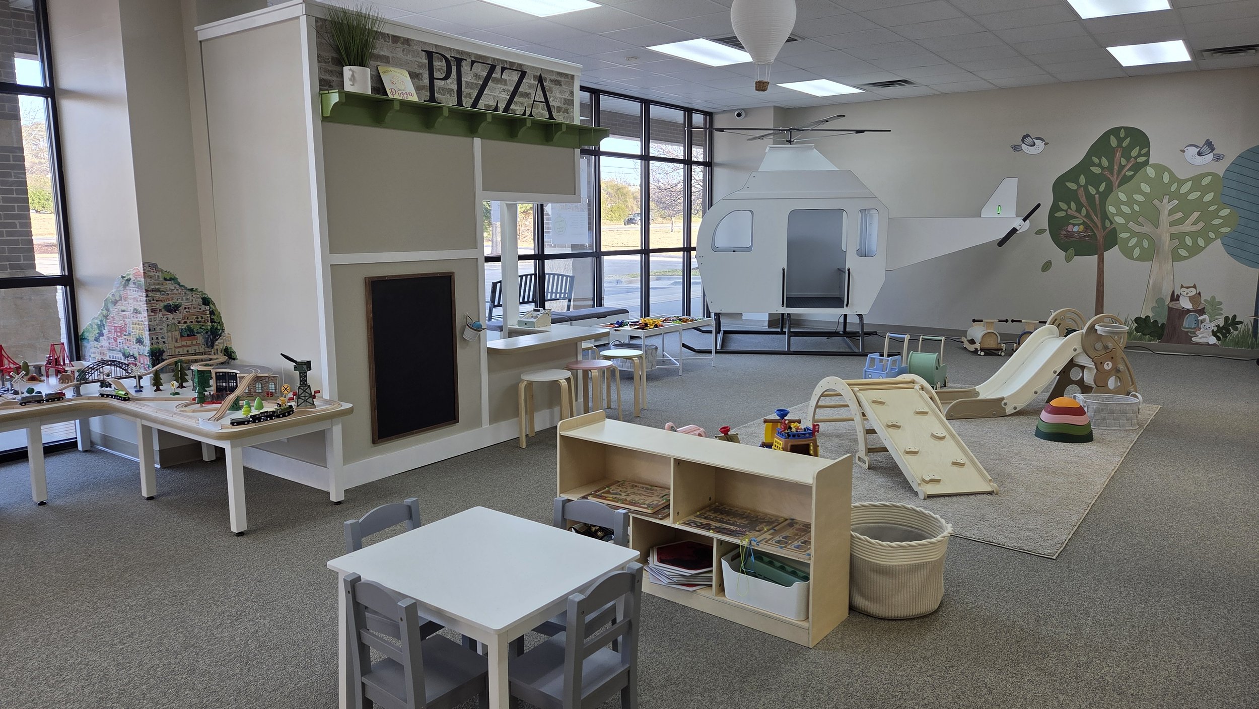 Indoor children's play area with toy train set, small table and chairs, playground slide, and a train-shaped hidden room, decorated with trees and animals on walls.