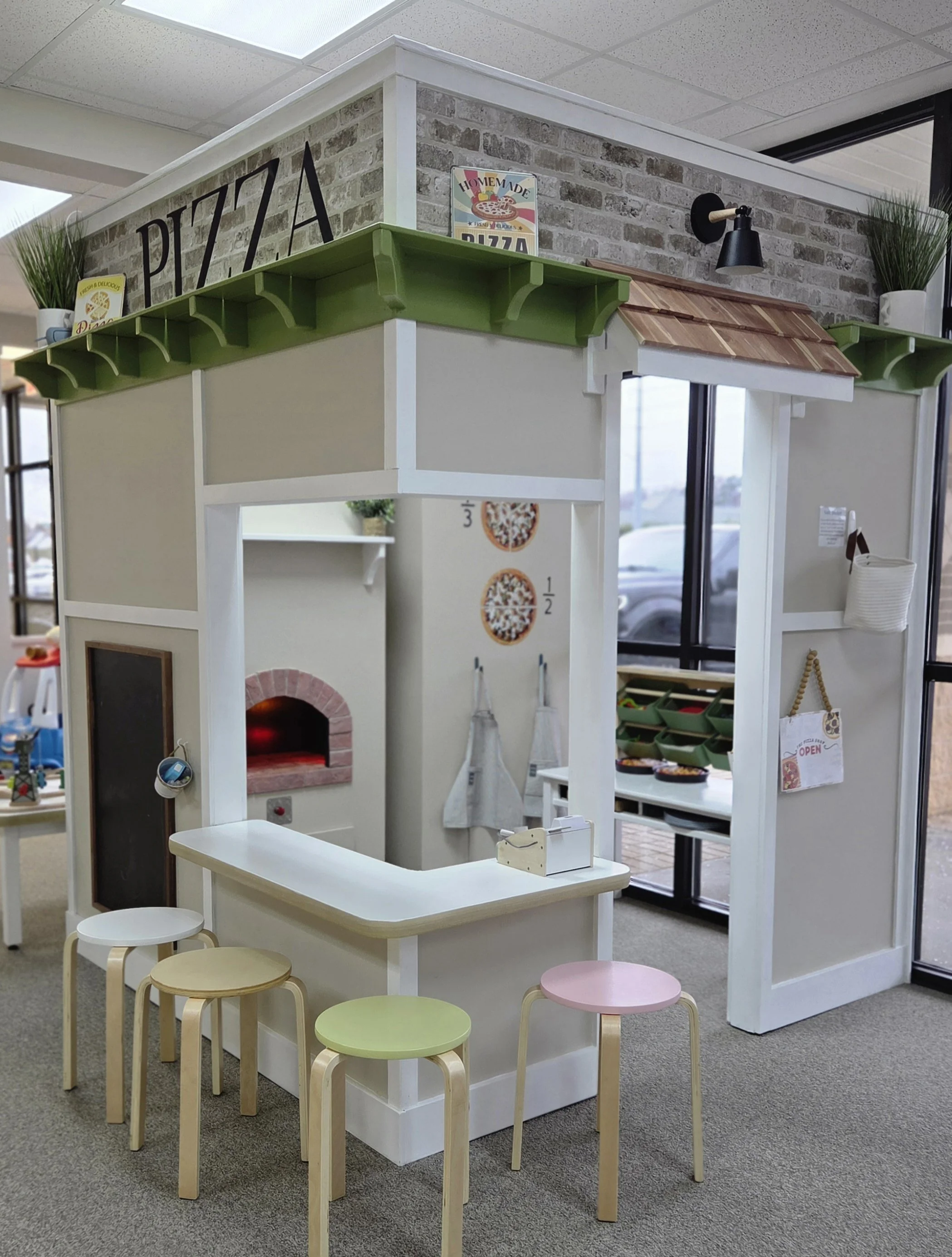 Miniature play kitchen designed as a pizza shop with a pretend oven, utensils, and counter, surrounded by small multicolored stools, inside a well-lit room with large windows.
