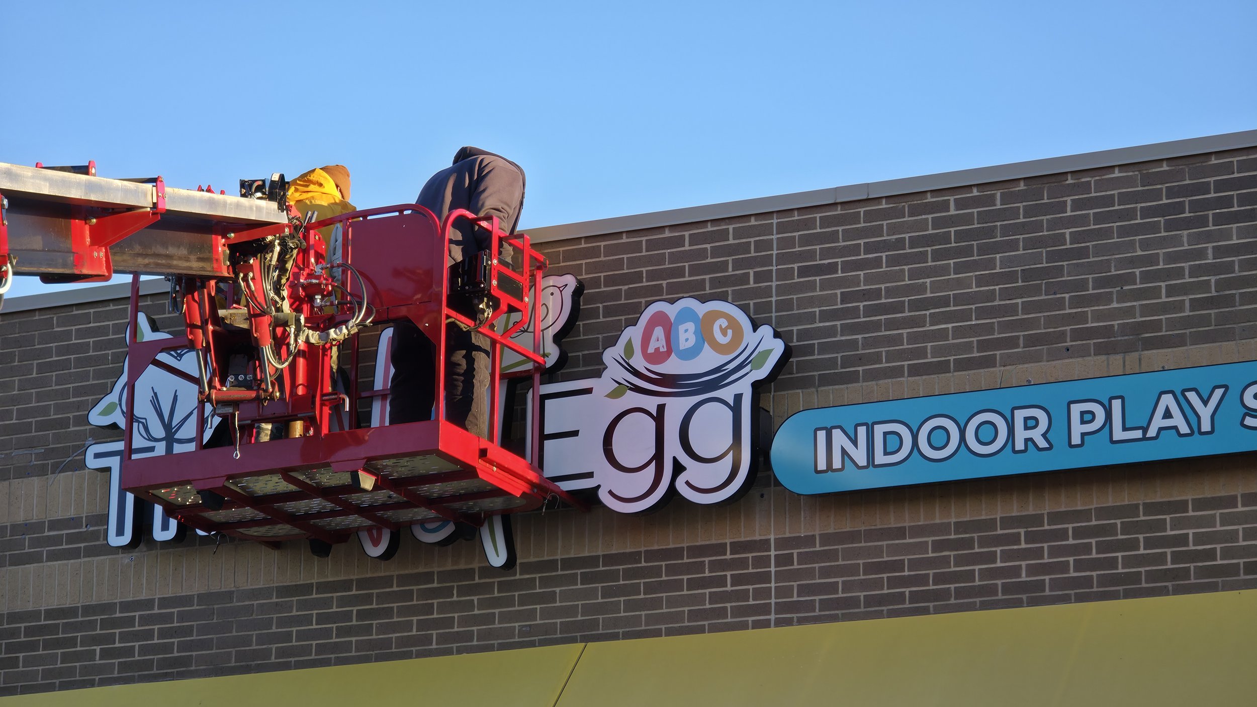 Worker on a red lift installing a sign that reads 'Egg Indoor Play Center' on a brick building.
