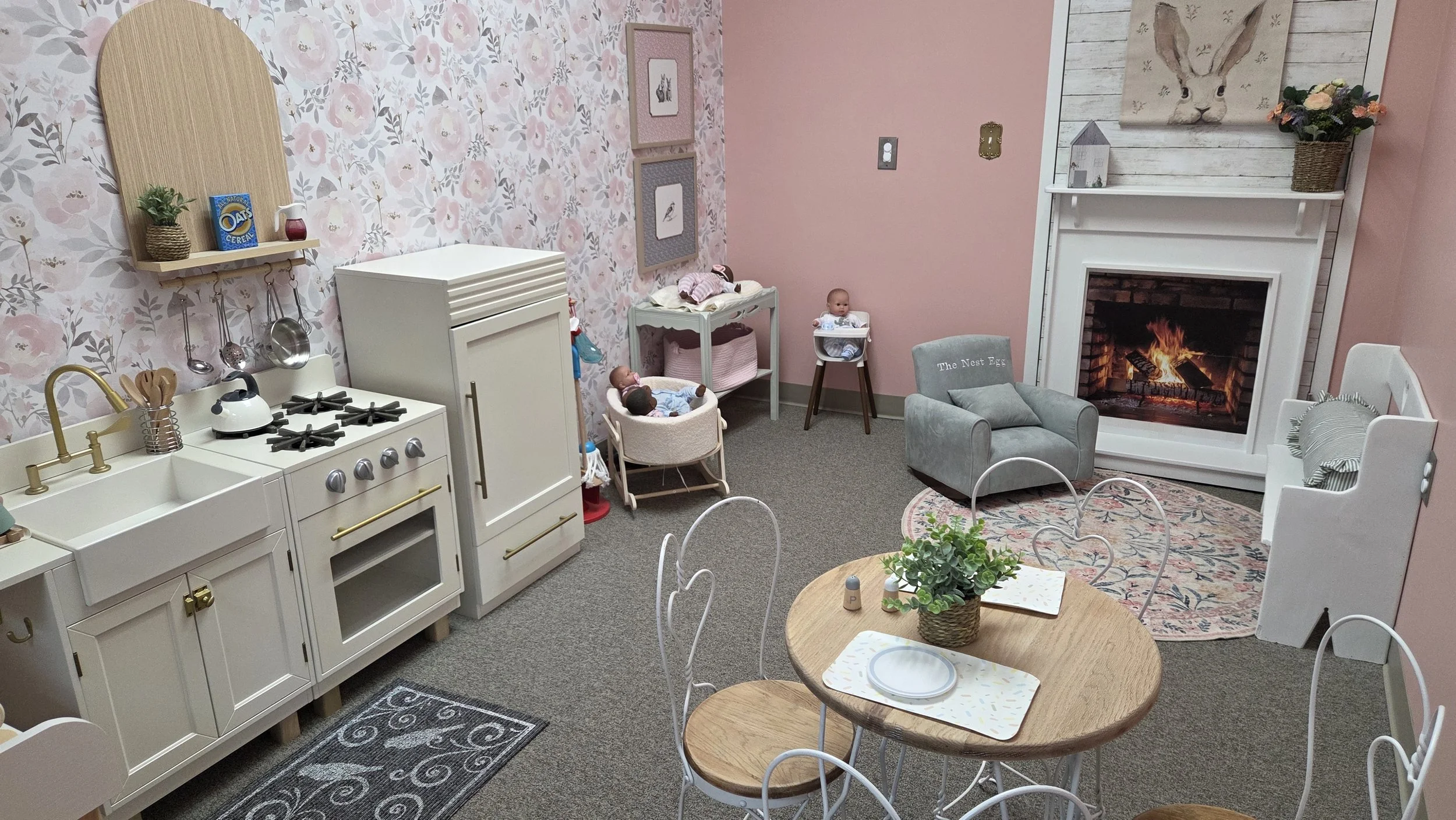 A cozy indoor space designed for children, featuring a play kitchen, dolls, a small family-sized table with chairs, a fireplace with a bunny painting above, and a plush armchair. The room has soft pink and floral wallpaper with decorations.