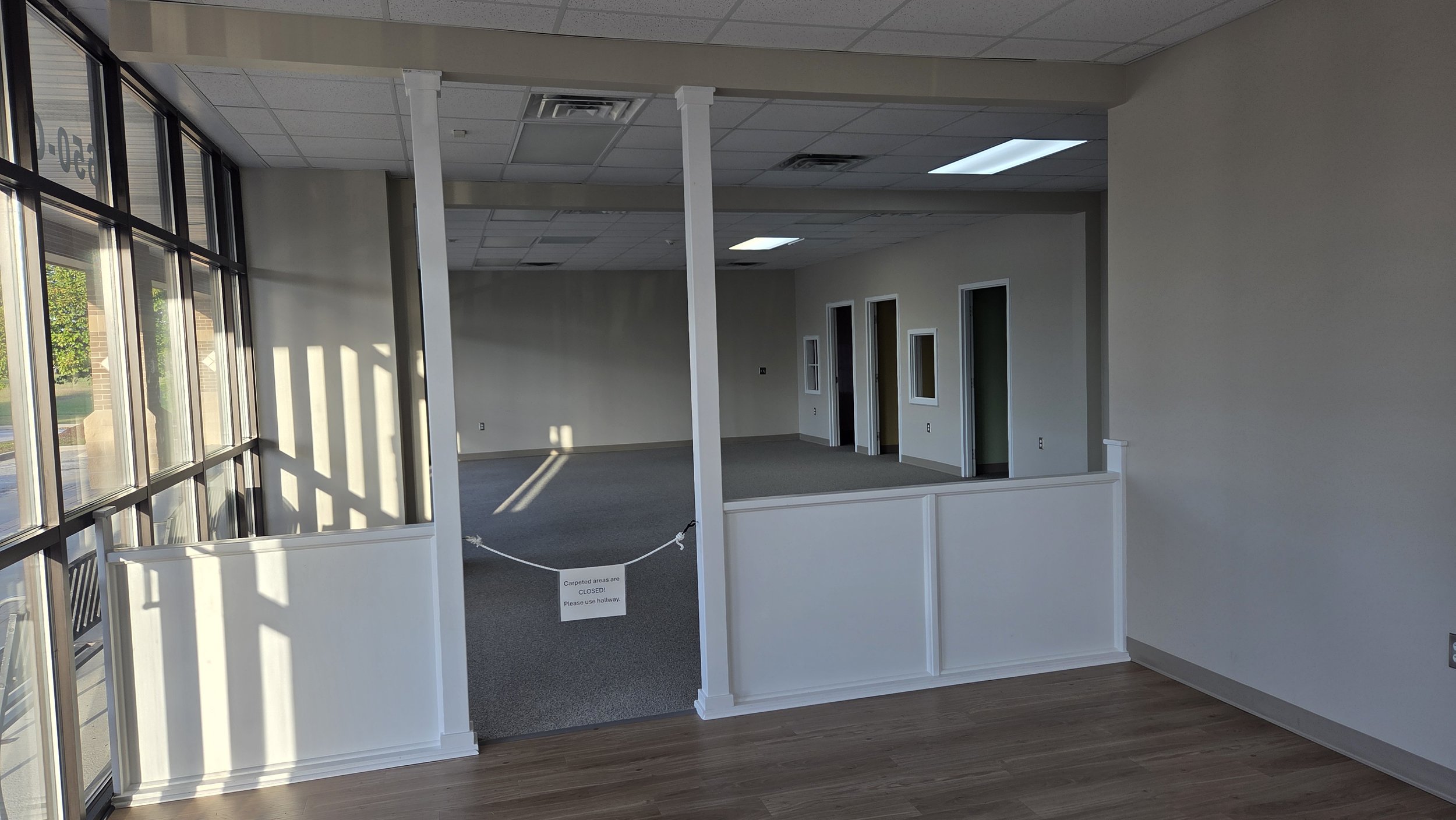 Empty office space with large window, Carpeted and hardwood flooring, Doorways, No furniture, Sunlight through window, Sign on rope barrier indicating carpeted areas are closed, Drop ceiling with fluorescent lights.