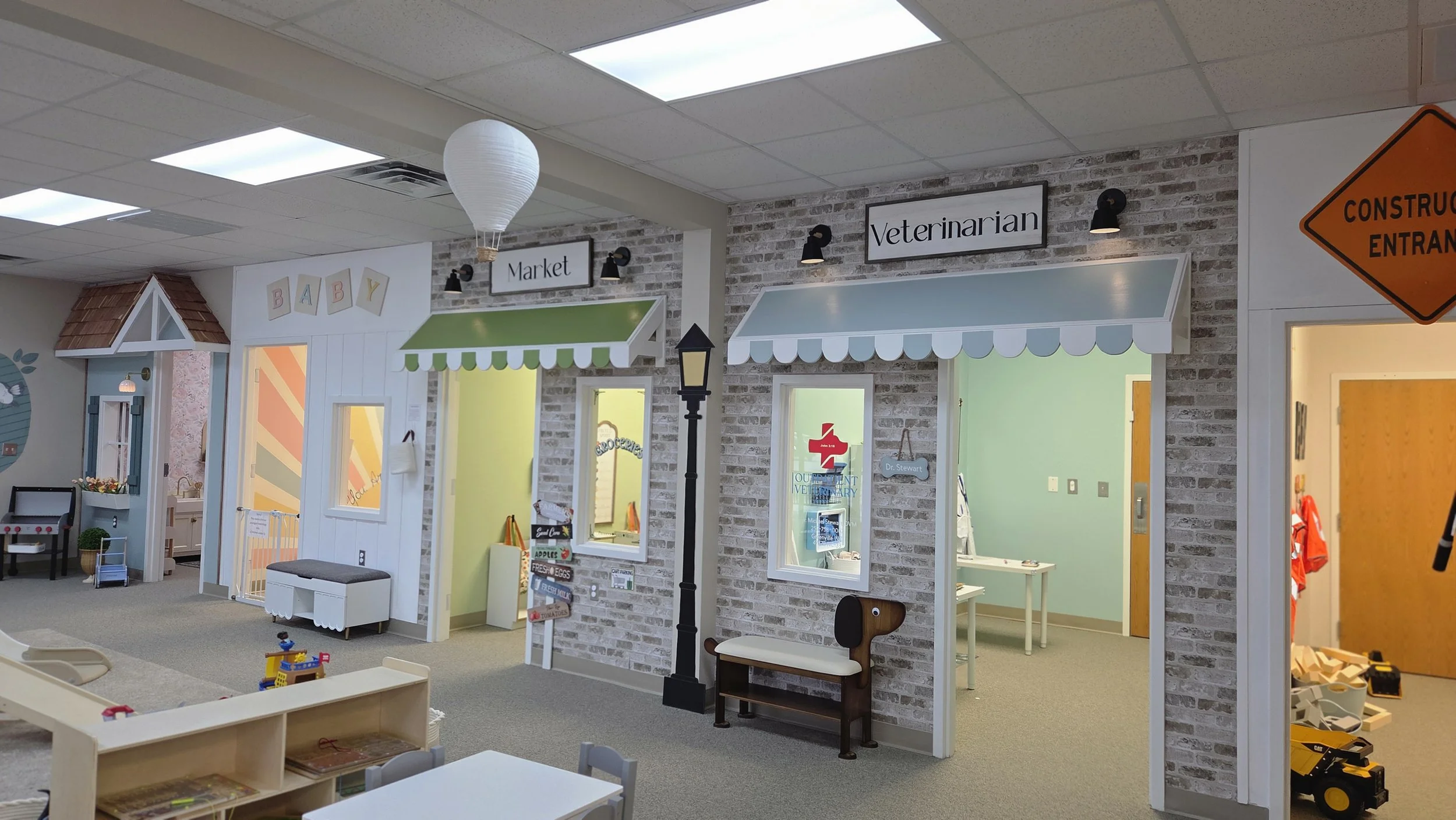 Children's play area with miniature storefronts labeled 'Market' and 'Veterinarian', designed for role-playing, with small furniture, toys, and colorful decor.
