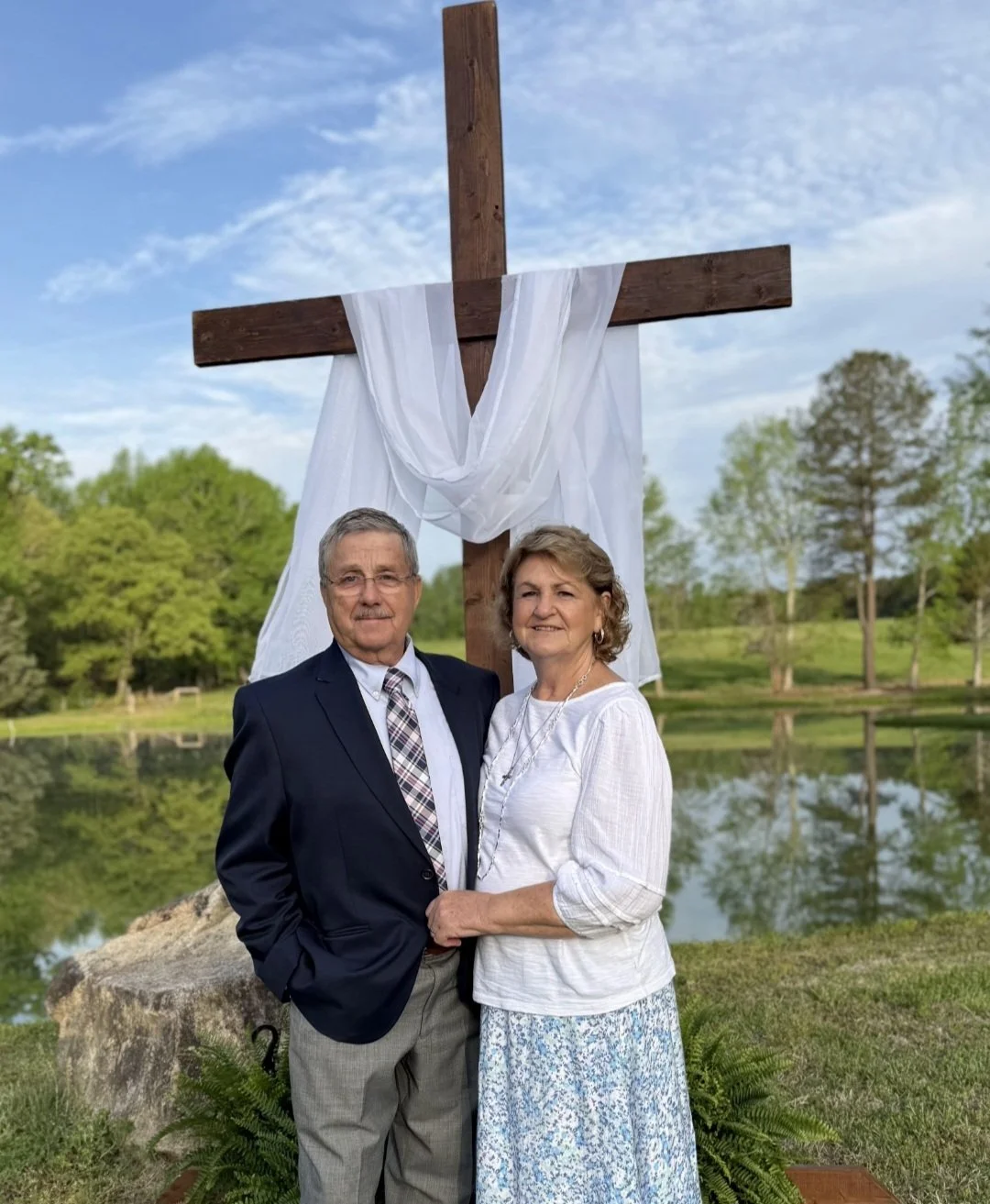 A middle-aged couple standing outdoors in front of a large wooden cross decorated with white fabric, beside a lake and green trees on a clear day.