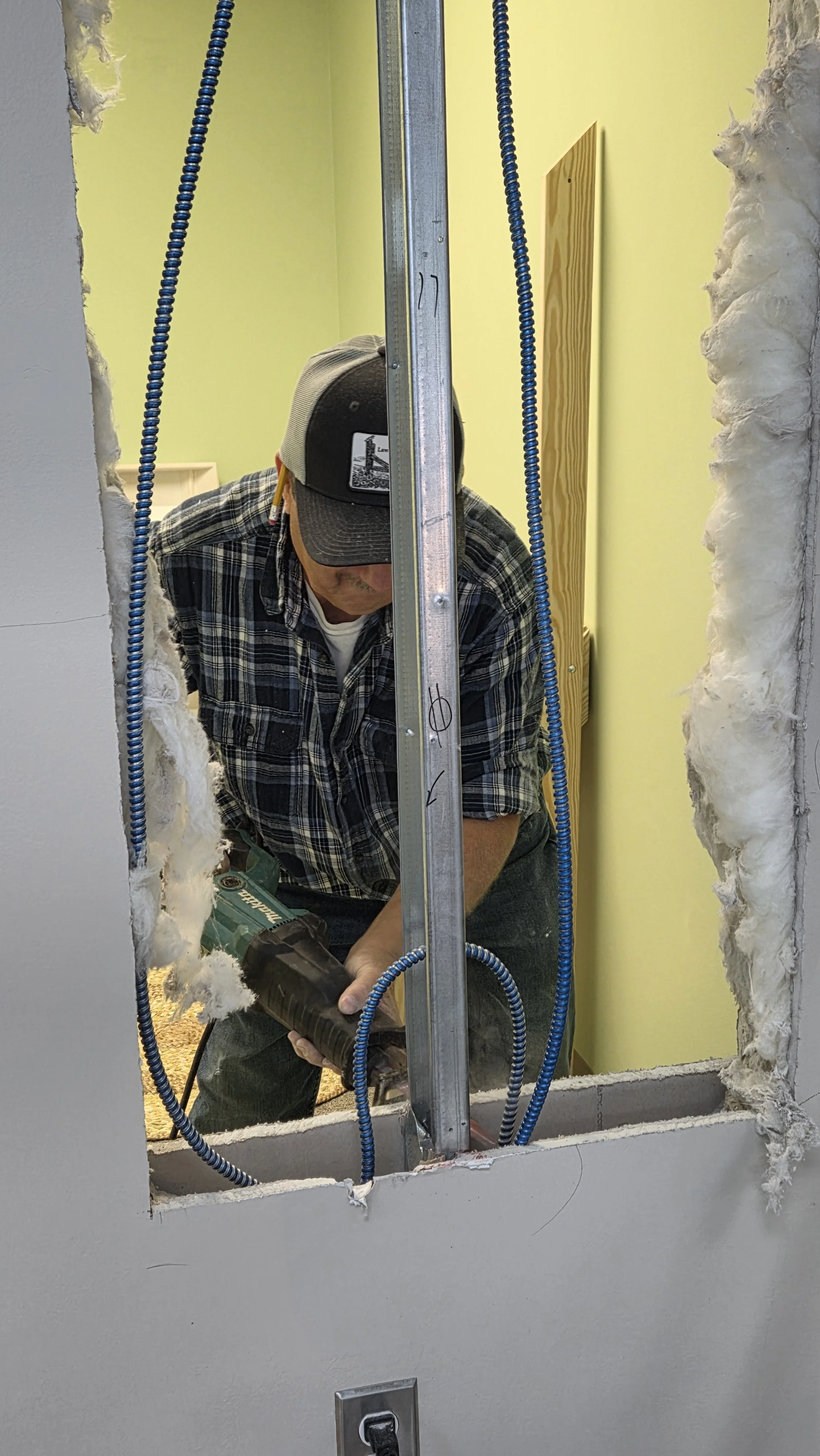 A person working on electrical wiring, using a cordless drill, seen through a hole in the drywall with blue electrical conduit and insulation surrounding the opening.