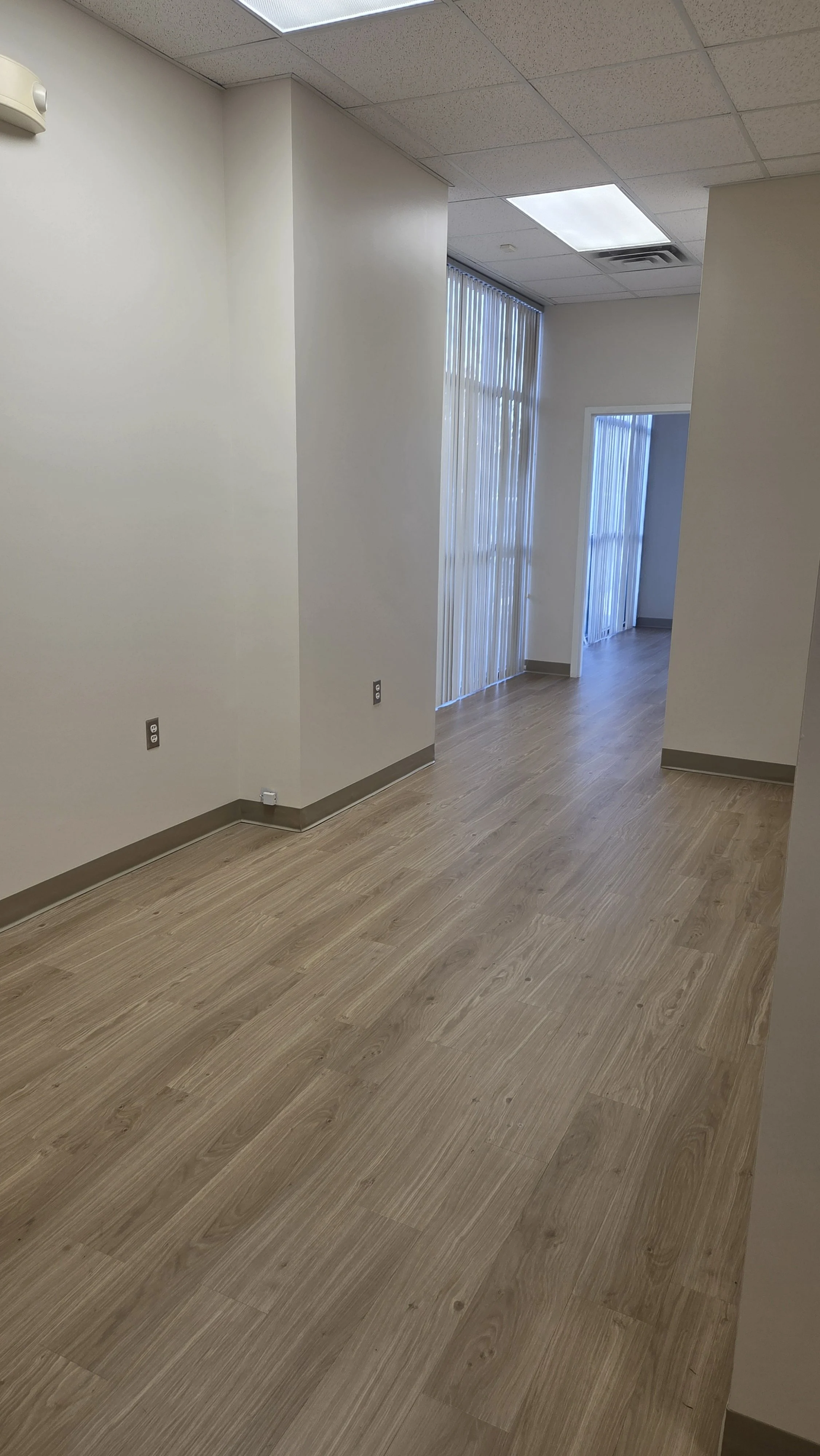 Empty office space with wood flooring, white walls, vertical blinds on windows, ceiling with rectangular fluorescent lights, and electrical outlets on the wall.