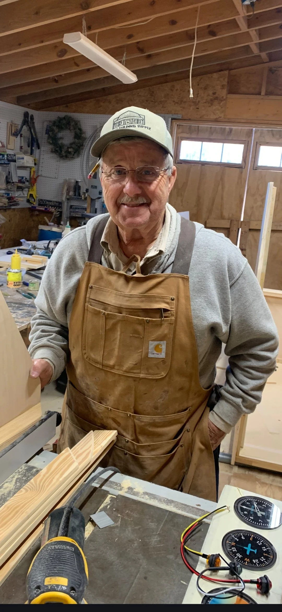 A smiling older man wearing glasses, a beige Carhartt apron, a gray hoodie, a cap, and standing in a woodworking shop filled with tools, wood pieces, and workbenches.