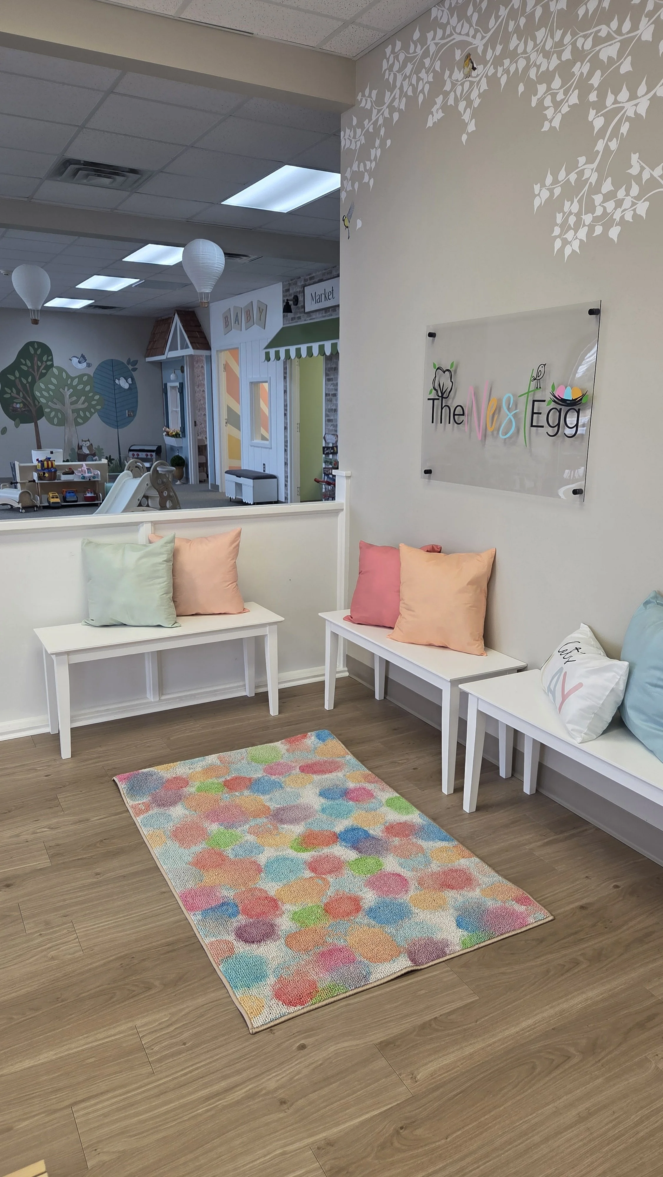 A cozy waiting area inside a children's store with white benches adorned with pastel-colored pillows, a colorful polka dot rug on wooden flooring, and a wall sign reading 'The Nest Egg' with playful lettering. The background features a play space with toys and decor resembling a small village with trees and houses.
