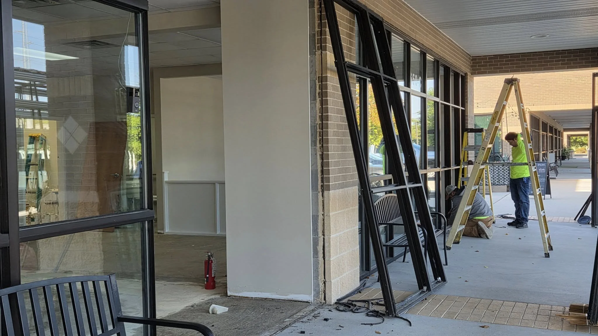 Construction workers installing or repairing large glass windows on the storefront of a commercial building. One worker is kneeling on the ground, and another is standing nearby, both engaged in work with tools. A yellow ladder is set up at the site.