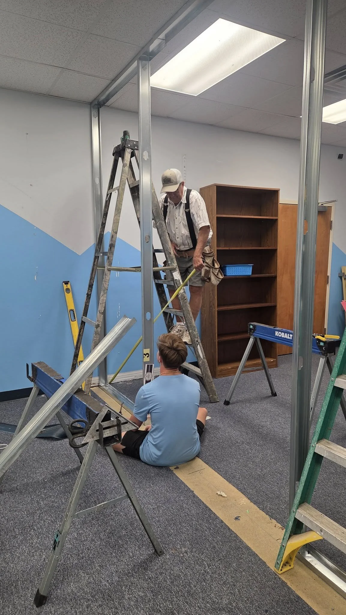 Two men are working on constructing a metal frame in a room with blue walls and a grey carpet. One man is sitting on the floor, and the other is standing on a ladder.