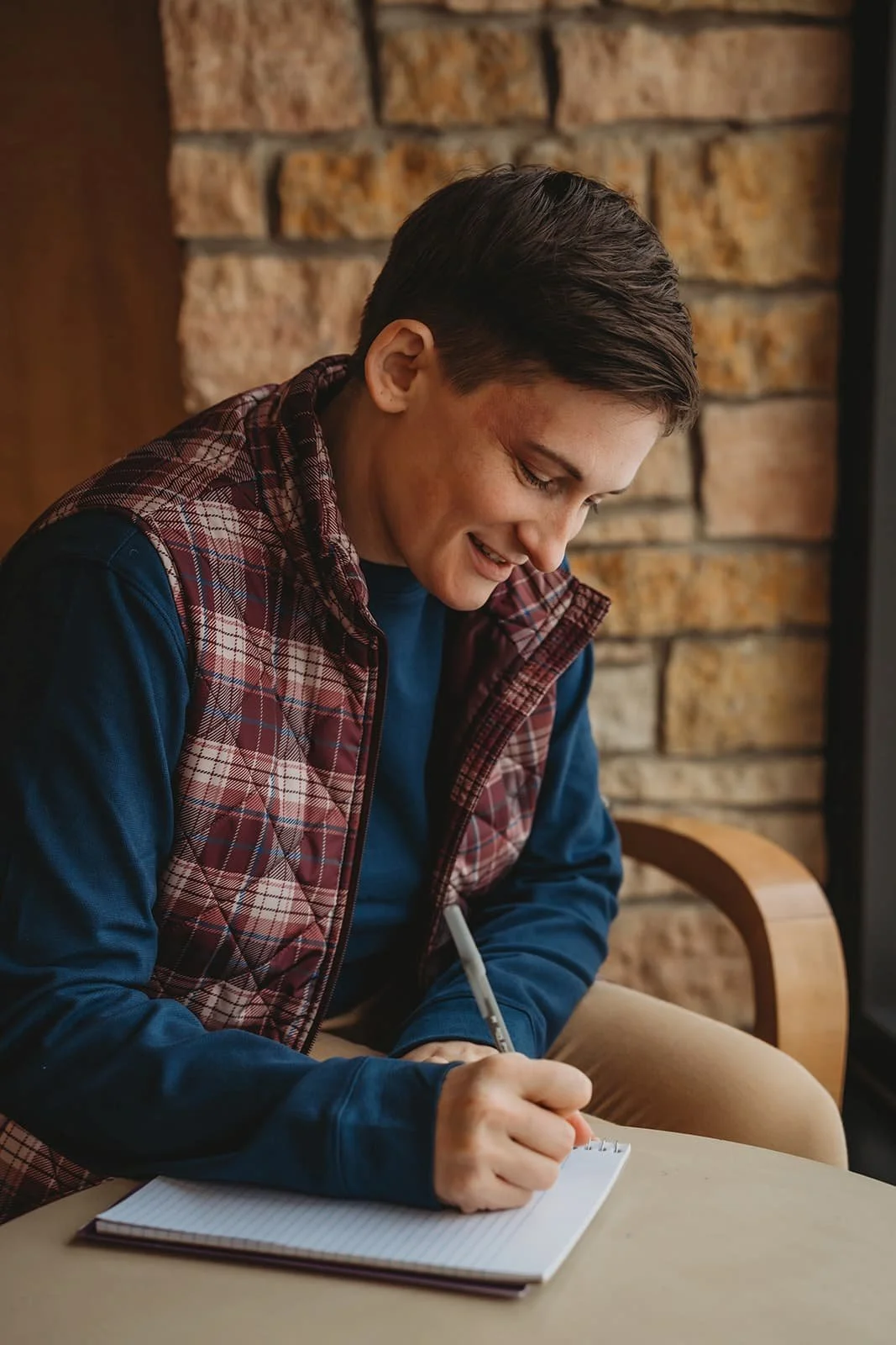 A young person sitting at a table, writing in a notepad, smiling, with a brick wall background.
