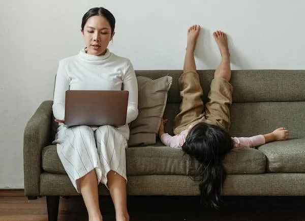 Person sitting on a couch working on a laptop while a child lies upside down with feet on the wall and head hanging off the couch.