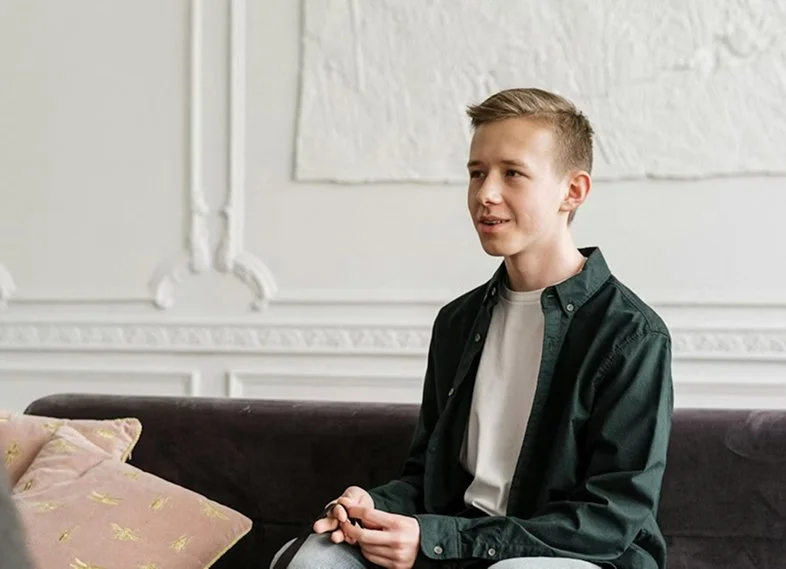 A young person sitting on a dark-colored couch in a well-lit room with decorative wall molding, holding an object in their hands and smiling slightly.