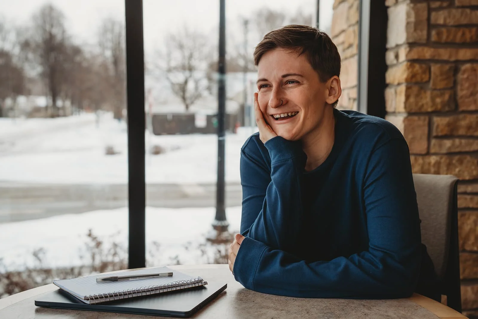 A person sitting at a table near a window during winter, smiling and resting their head on their hand, with a notebook, pen, and laptop on the table.
