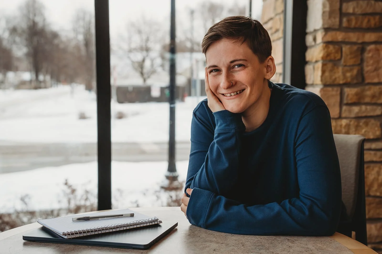 A young person with short hair and a nose piercing sitting at a table, smiling and resting their head on their hand, with a notebook and pen in front of them, inside a library with a large window showing a wintery outdoor scene.