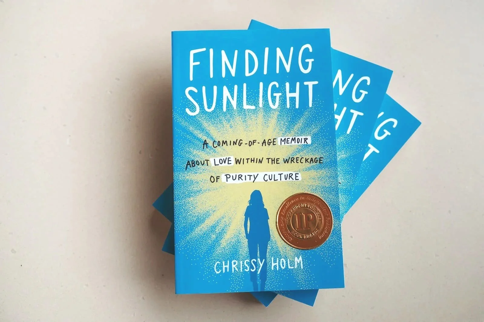 Stack of three copies of the book titled 'Finding Sunlight' by Chrissy Holm, with a blue cover featuring a silhouette of a person walking towards a bright burst of sunlight, and a round, bronze-colored award sticker on the front.