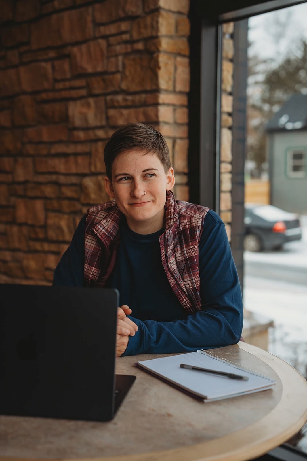 A young person with short hair, wearing a plaid vest, sitting at a round table with a laptop, notebook, and pen, looking out the window with a slight smile.