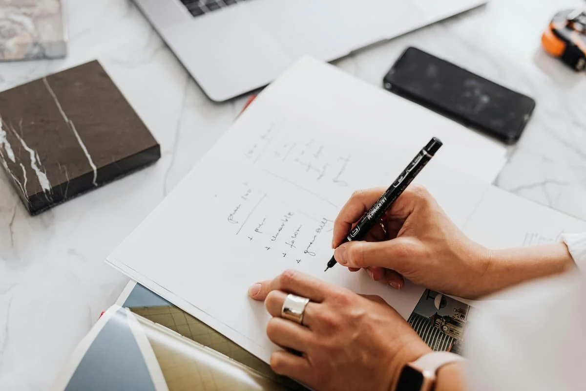 Person writing notes or sketches on a white paper with a black marker, surrounded by a laptop and smartphone.