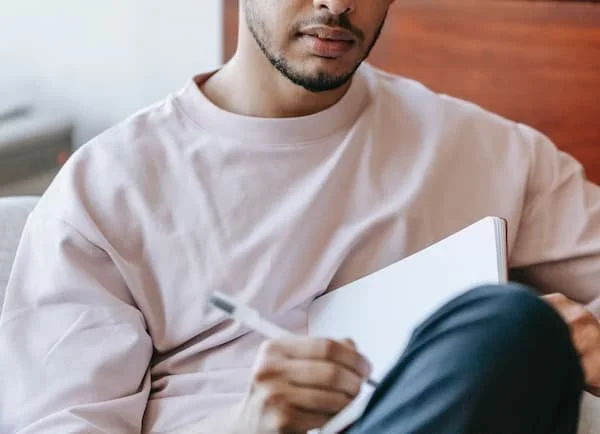 A person in a white T-shirt sitting on a couch, holding a notebook and a pen.