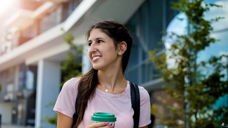 Young person with long brown hair holding a coffee cup, standing outdoors in an urban setting with modern buildings and green trees.