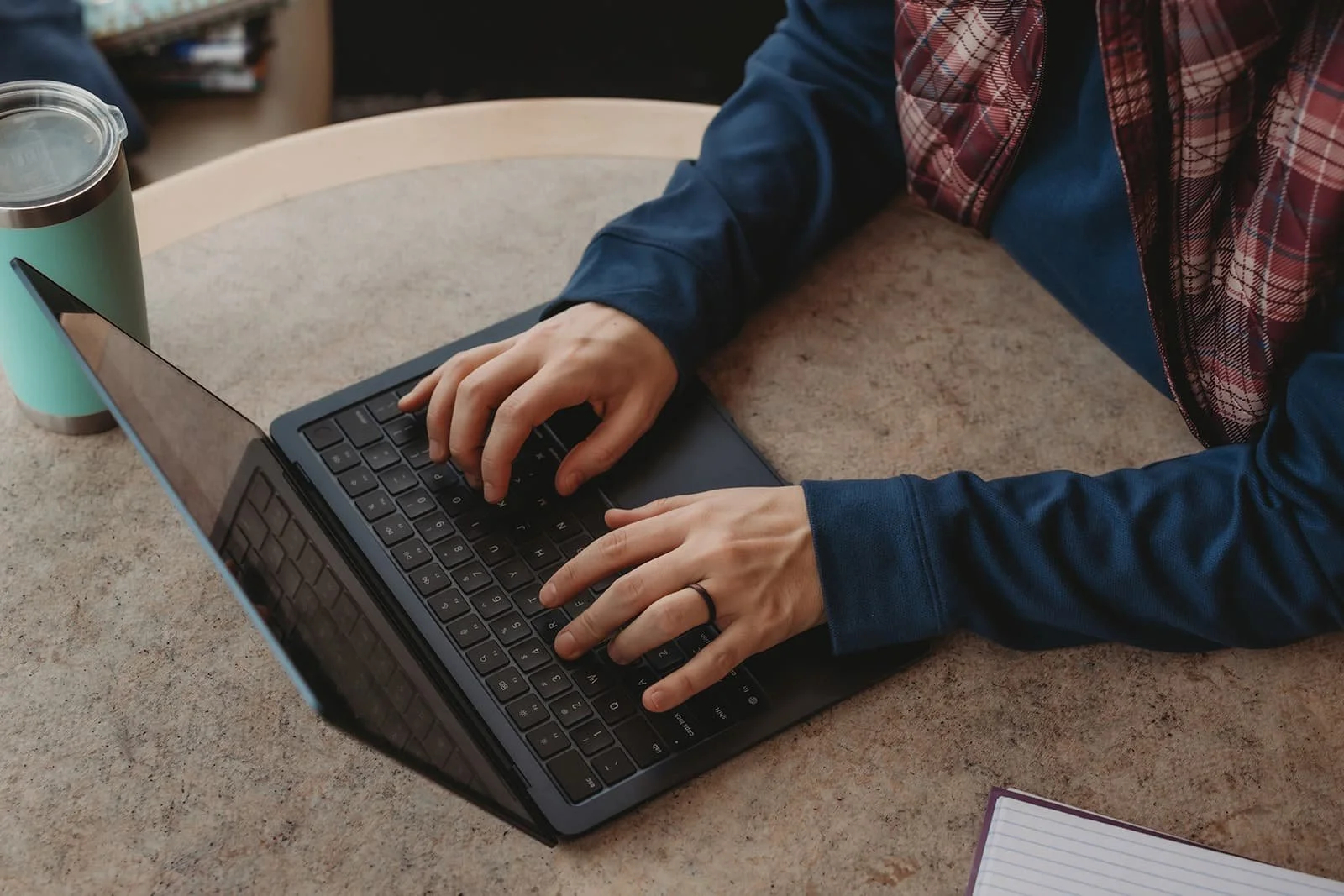 Person using a laptop computer at a table with a teal travel mug and a notepad nearby.