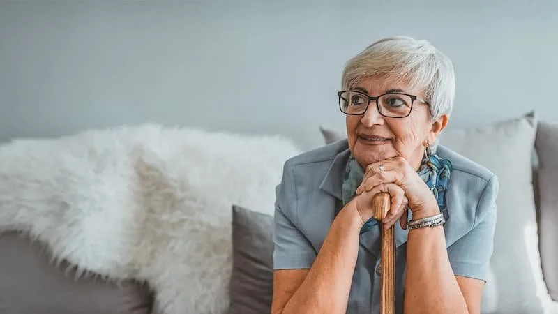 An older adult with short gray hair and glasses, smiling and resting their chin on a cane, sitting on a couch with pillows and a fluffy white blanket behind them