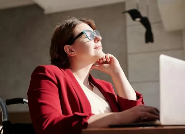 Person with glasses wearing a red blazer sitting at a desk with a laptop, looking up thoughtfully in a well-lit room.
