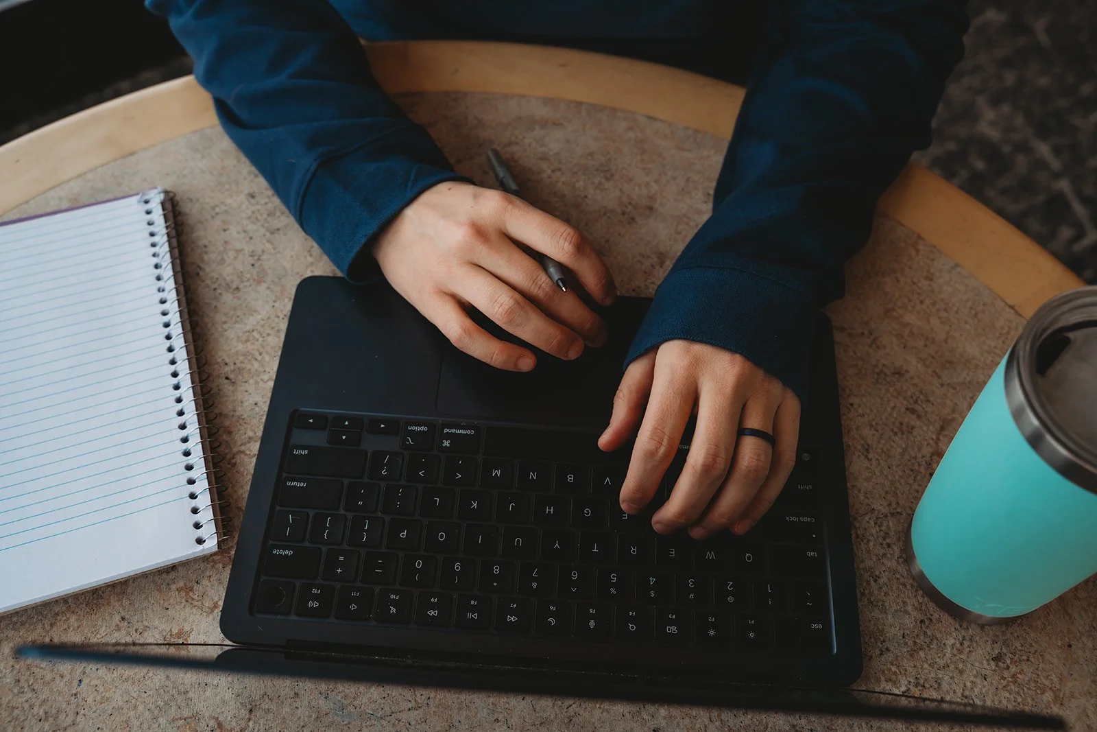 Person using a black keyboard with a notebook and a teal travel mug on a round wooden table.