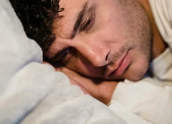 A person with curly dark hair lying on a pillow with his eyes closed, appearing to be sleeping or resting.