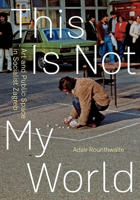 A person drawing on the street with chalk in front of a green car and a group of people standing outside a building in the background.