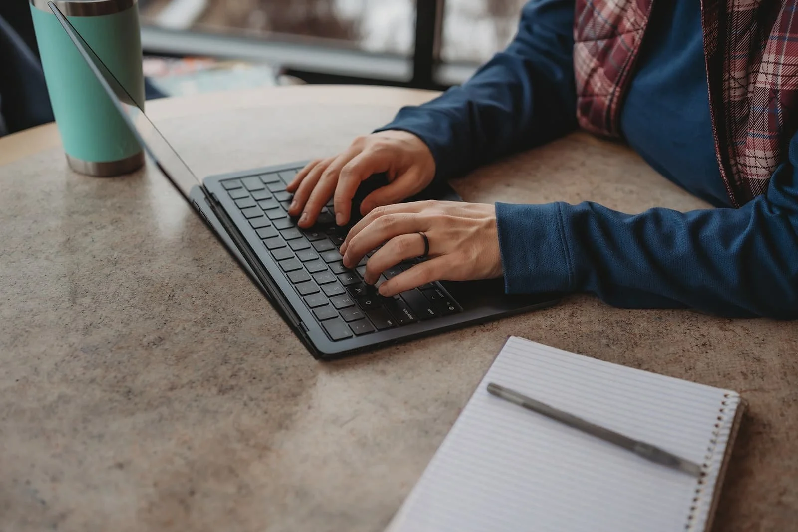Person typing on a laptop keyboard at a table with a notebook and pen nearby.