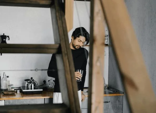A person with dark hair and a beard holding a glass of drink, standing in a modern kitchen, seen through a wooden ladder or structure.