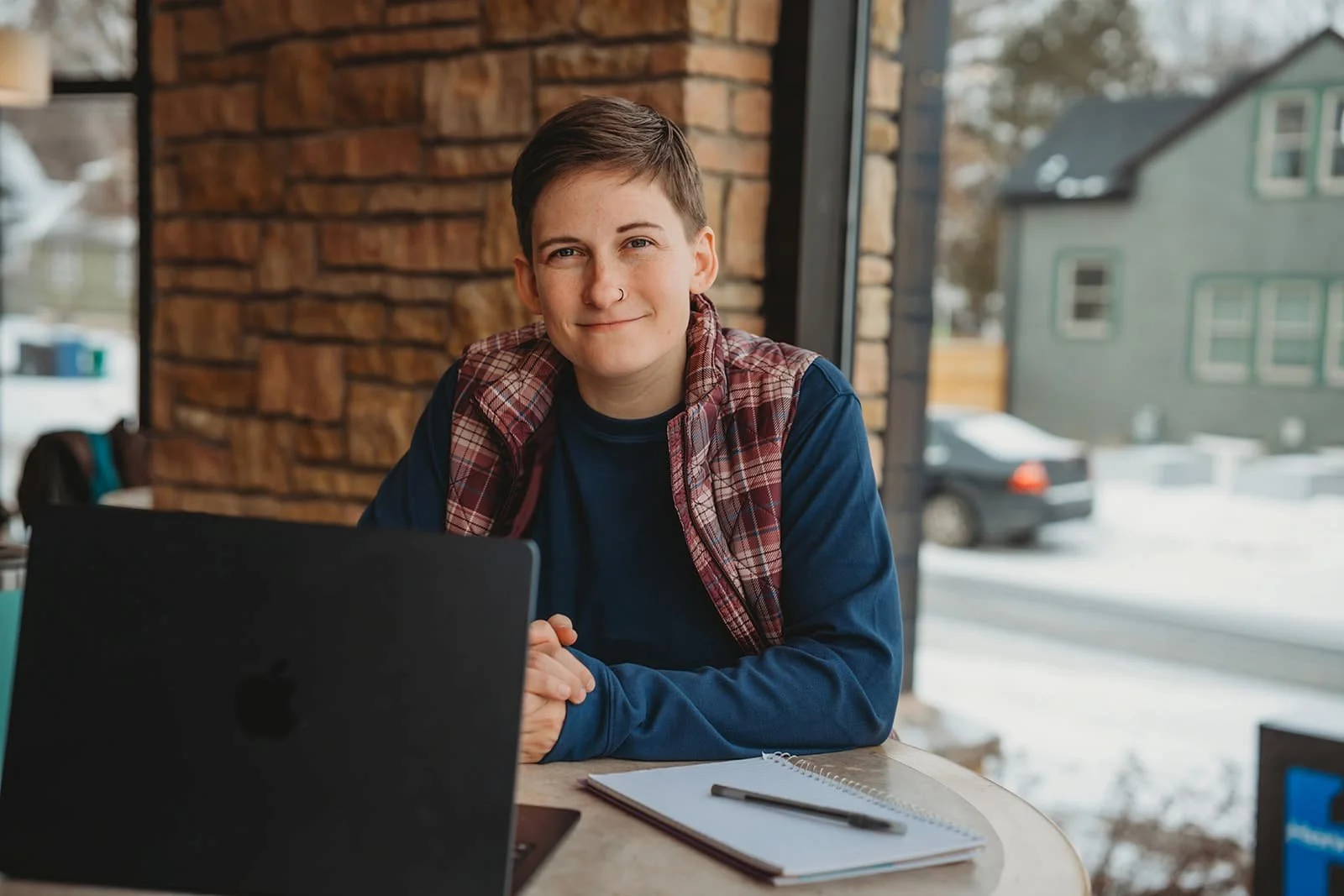 Young person with short hair, wearing a blue shirt and plaid vest, sitting at a table with a laptop, notebook, and pen, inside a library during winter.