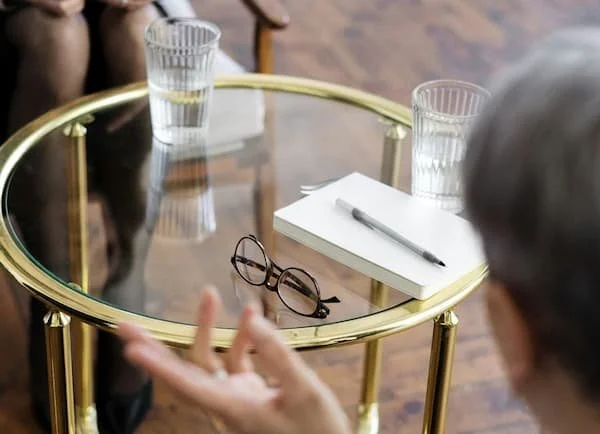 A round glass-top table with a gold frame, two glasses of water, a notebook, a pen, and a pair of glasses, with a person making a hand gesture nearby.