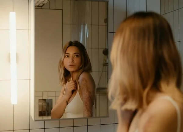 A person with shoulder-length blonde hair looking at themself in a bathroom mirror with white tiled walls.