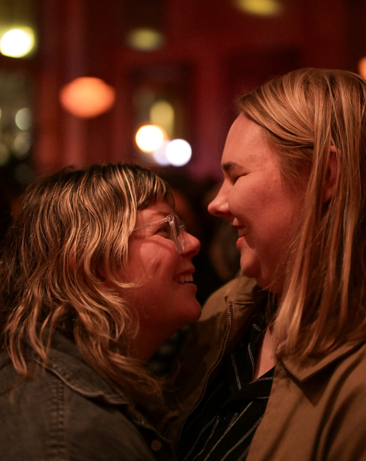 Two people with shoulder length blond hair are pictured smiling at each other.