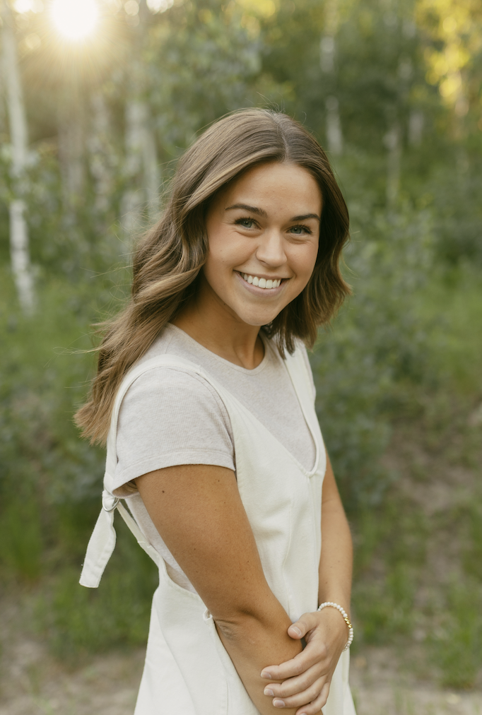 A young woman with long brown hair smiling outdoors in a wooded area during sunset, wearing a light-colored dress and bracelet.