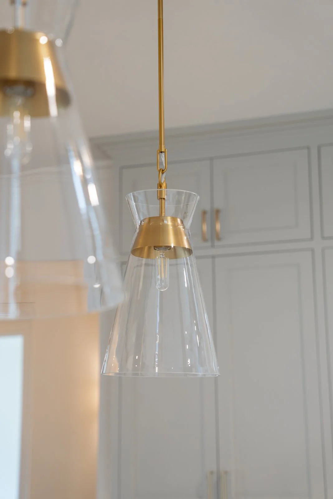 Close-up of a modern hanging glass pendant light with a gold fixture in a kitchen with white cabinets.