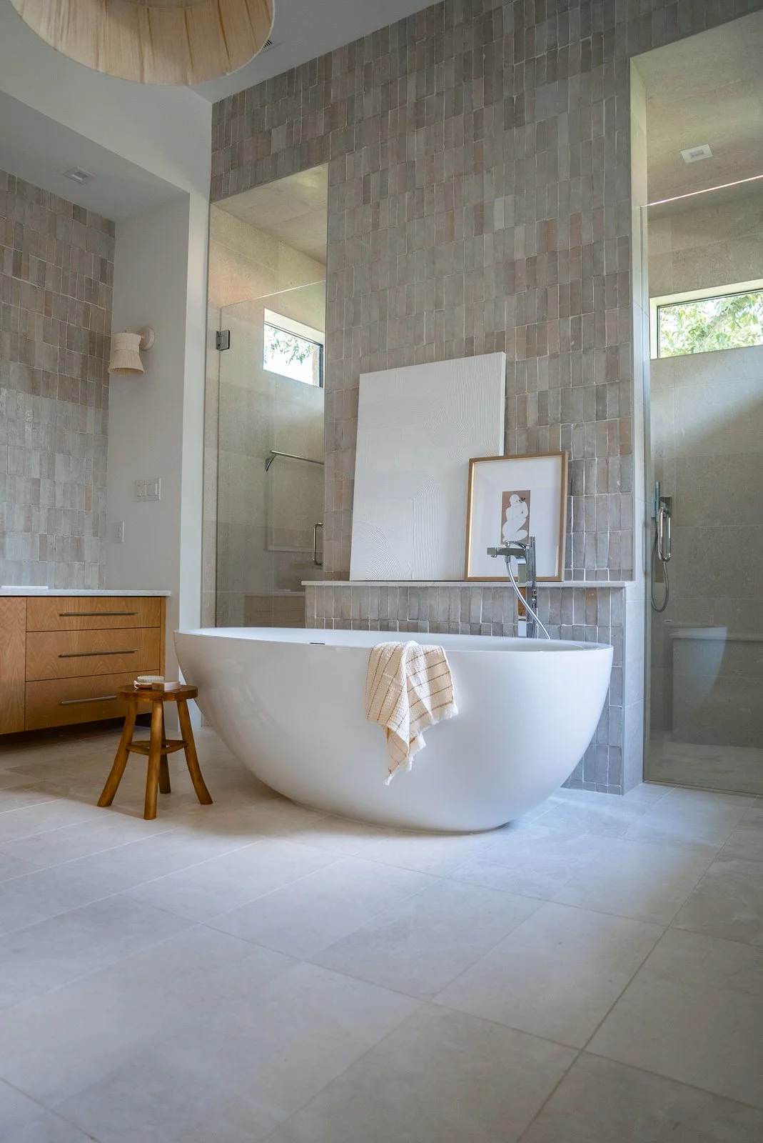 Modern bathroom with a white soaking tub, beige tiled floors, a small wooden stool with a towel, a wooden cabinet, framed artwork, and a glass-enclosed shower area.