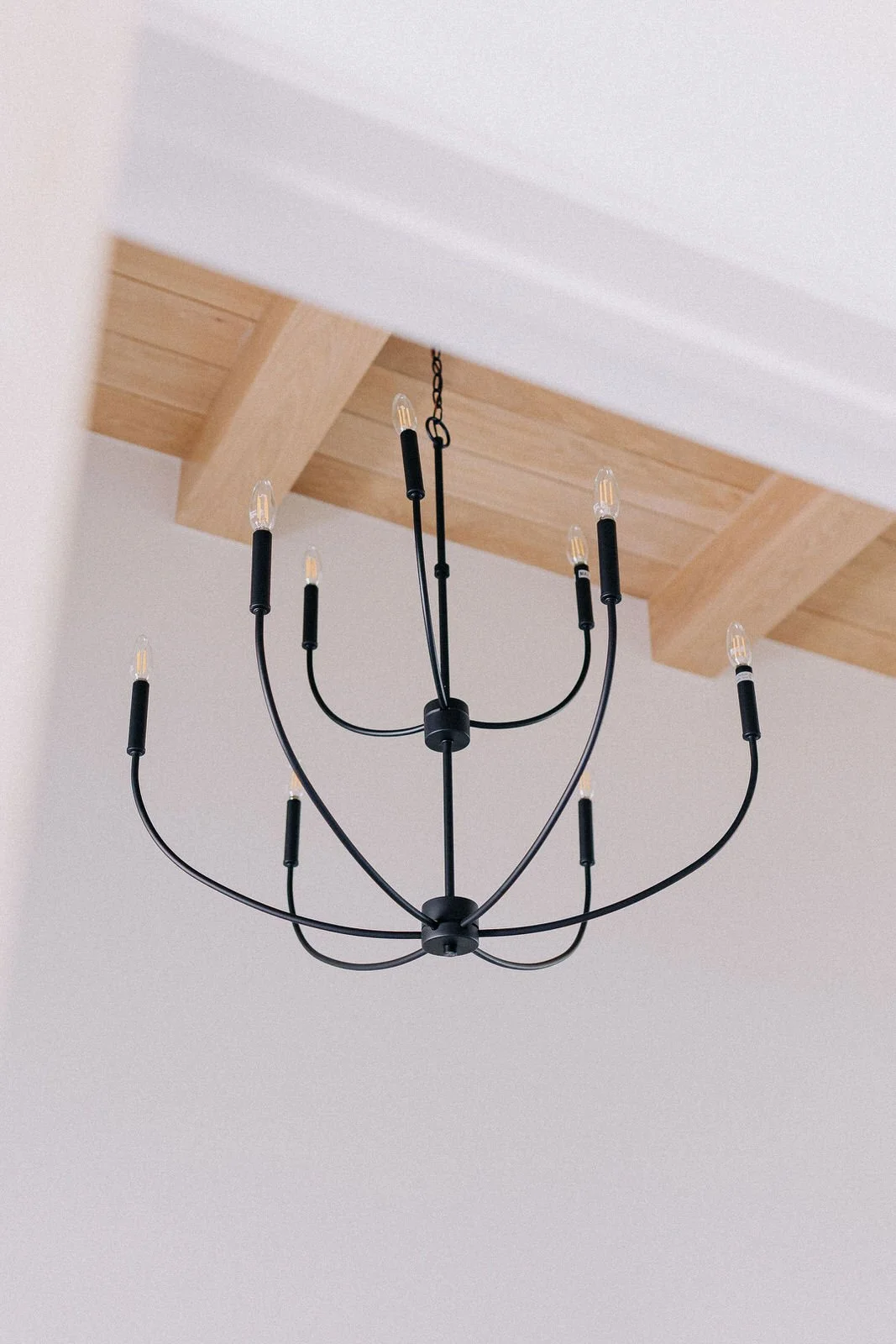 A modern black chandelier with multiple arms and small candle-like light bulbs hanging from a wooden ceiling beam.