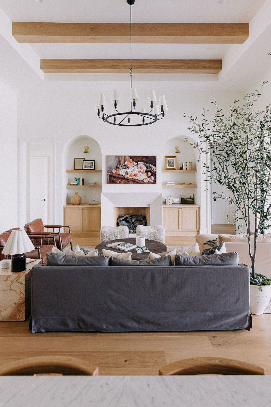 Living room with a black chandelier, white walls, wooden beams on the ceiling, a fireplace in the center, a large potted plant on the right, and various furniture including a gray couch and two armchairs.