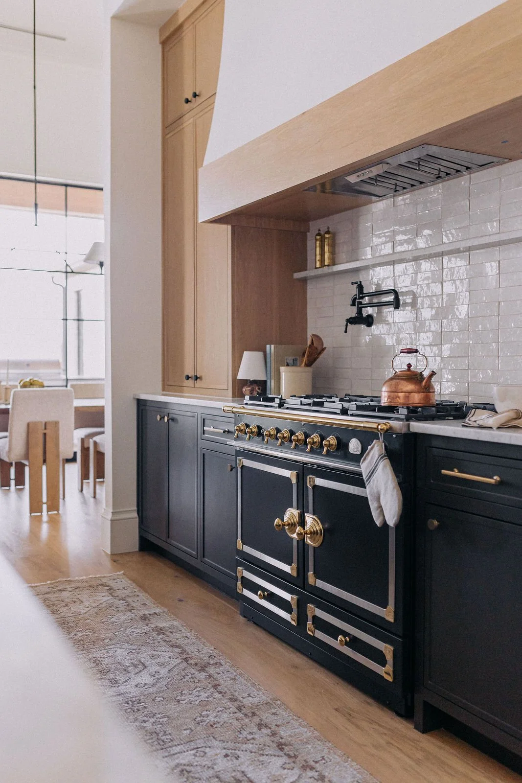 A modern kitchen with a black stove with gold accents, a copper kettle, wooden cabinets, and a white tiled backsplash.