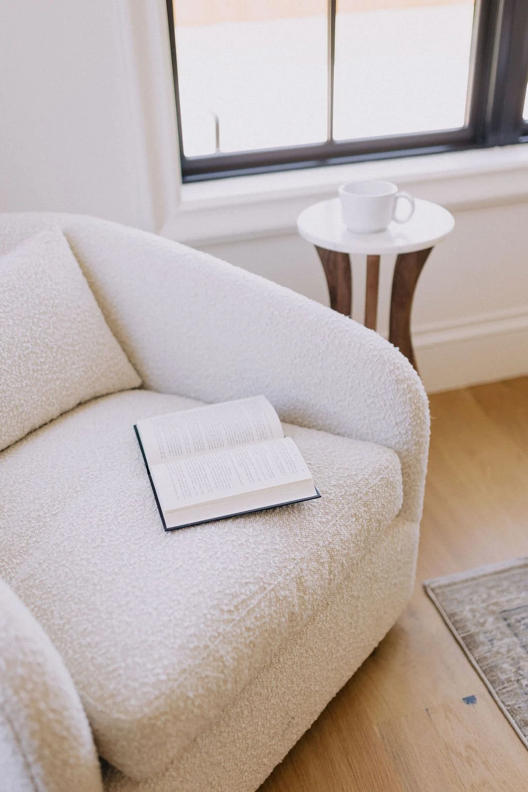 A cozy living room corner with a cream textured sofa, an open book resting on it, a small side table with a white cup, near a window with sunlight streaming in.